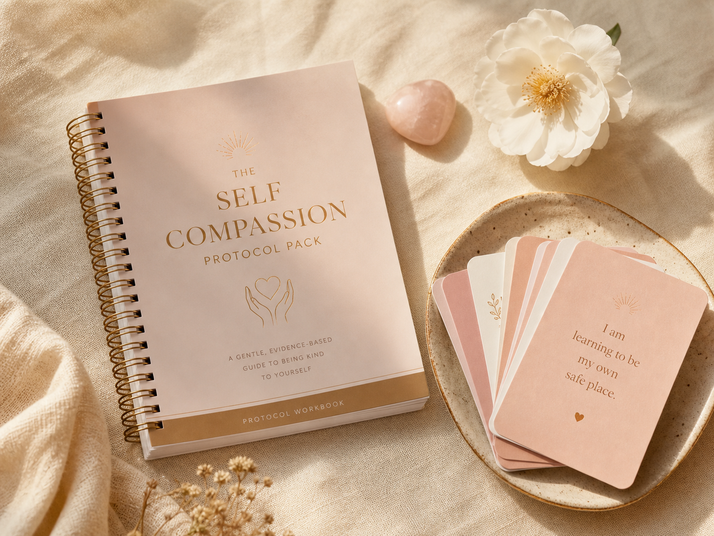 Soft flat lay of spiral workbook, affirmation cards, white flower and polished stone representing The Self Compassion Protocol Pack