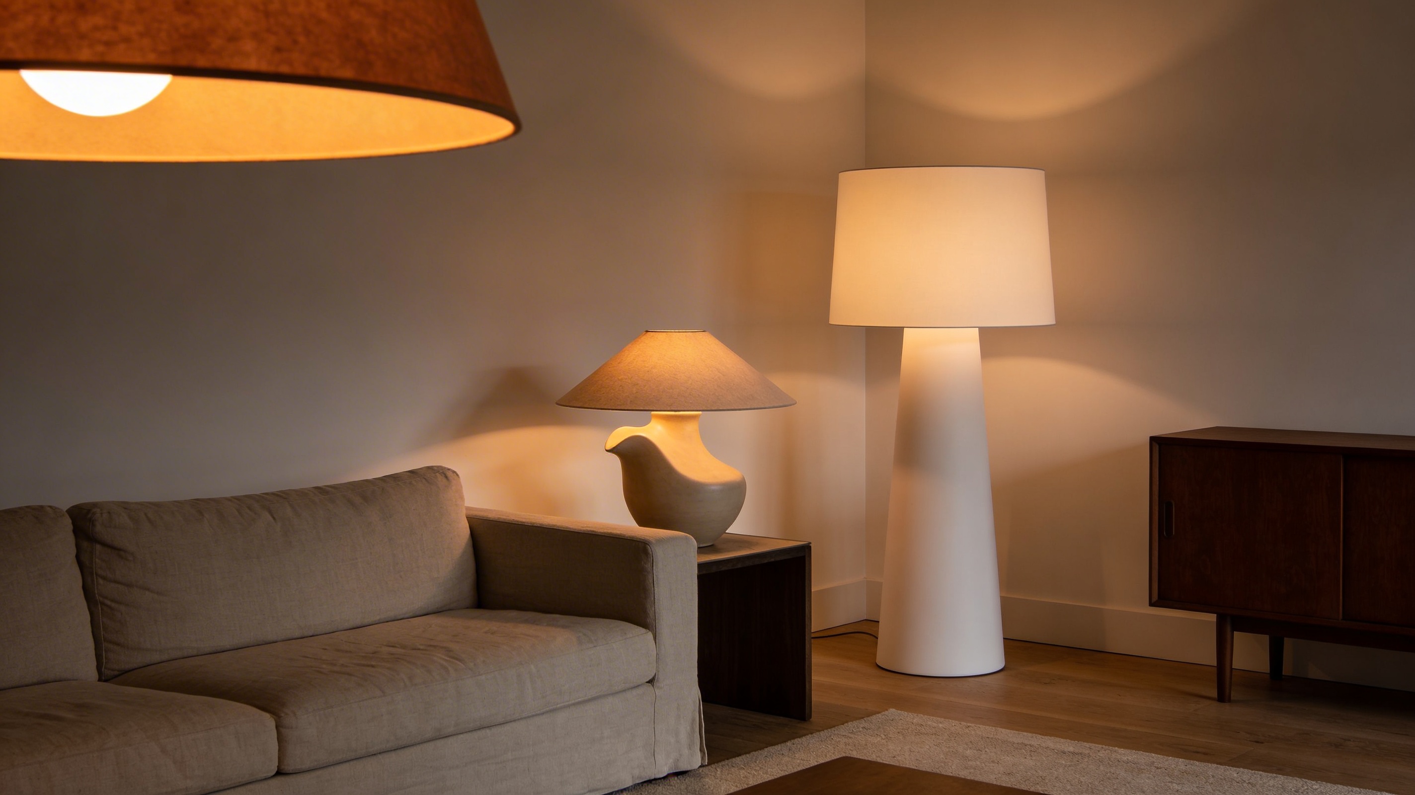 A beautifully layered modern American living room in 2026 with sculptural floor lamp, ceramic table lamp, and warm dimmed ceiling pendant — no exposed bulbs