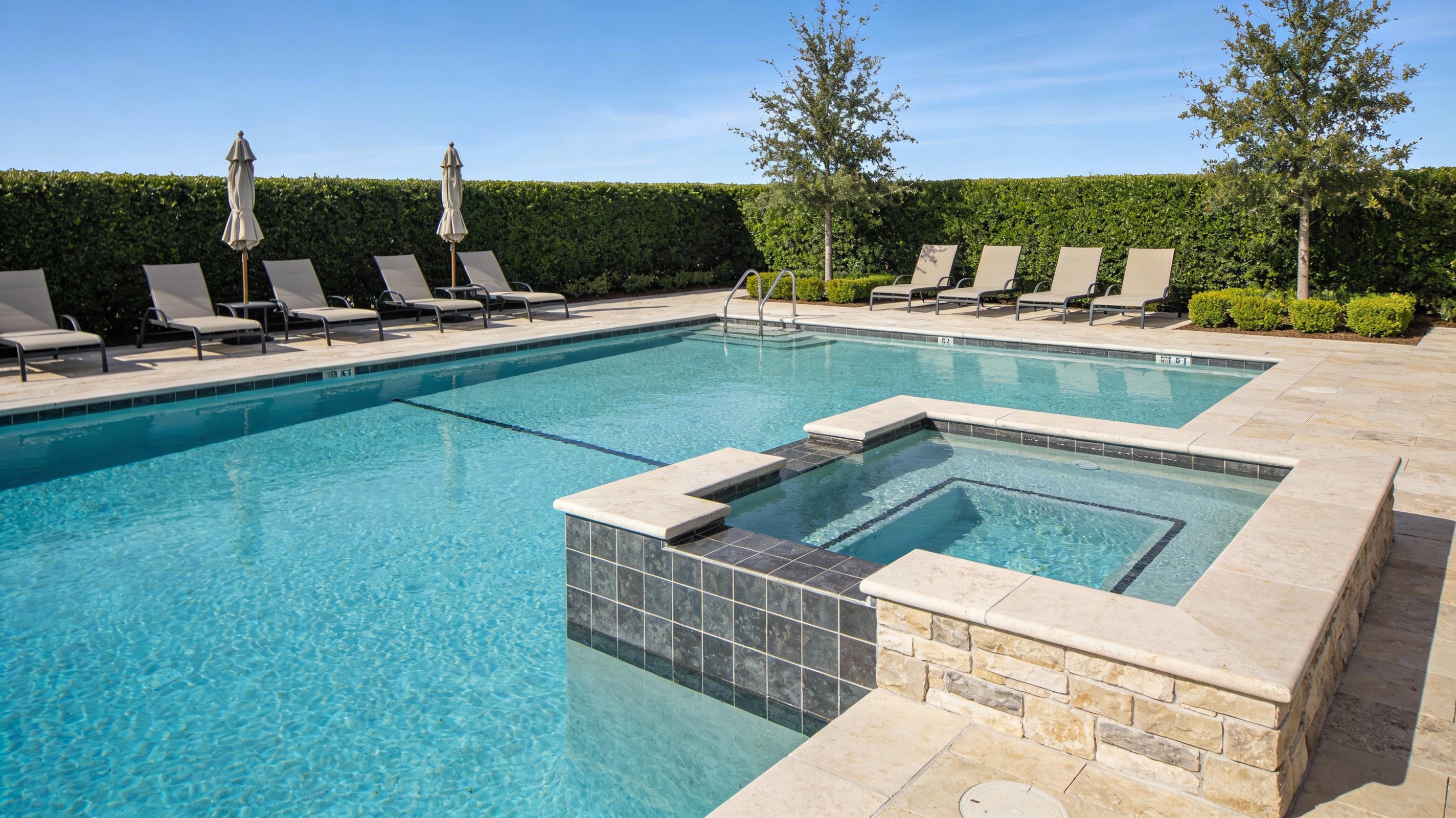 A view of an upscale commercial outdoor pool area at a modern hotel or apartment community, featuring a large rectangular blue pool with light stone coping, an attached raised spa, contemporary lounge chairs with umbrellas, and manicured greenery under a bright daytime sky.